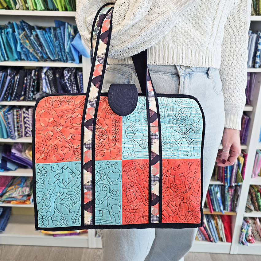 Person holding a colorful quilted craft tote bag with a pattern of geometric shapes in front of a fabric shelf