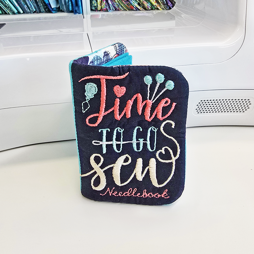 Embroidered needlebook with colorful text leaning against sewing machine