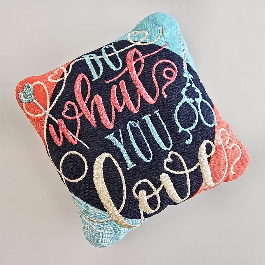 Decorative pin cushion with colorful text and heart designs on a light gray background