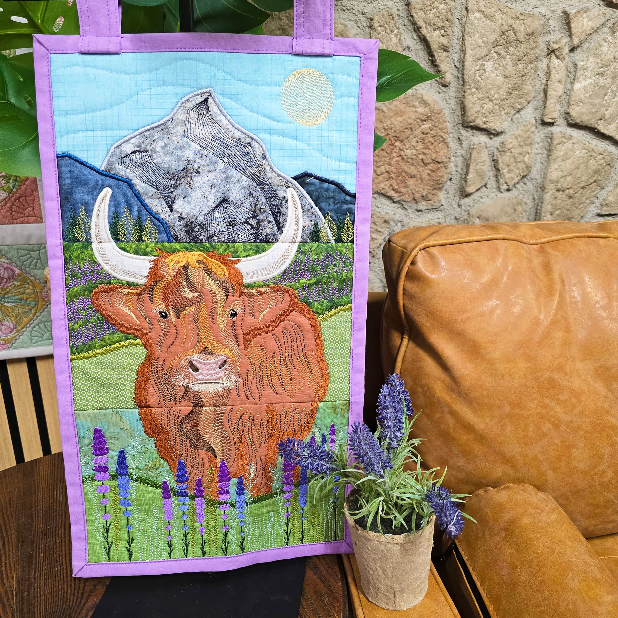 A handmade embroidered wall hanging featuring a Highland cow in a field with purple flowers in the sweet pea studio