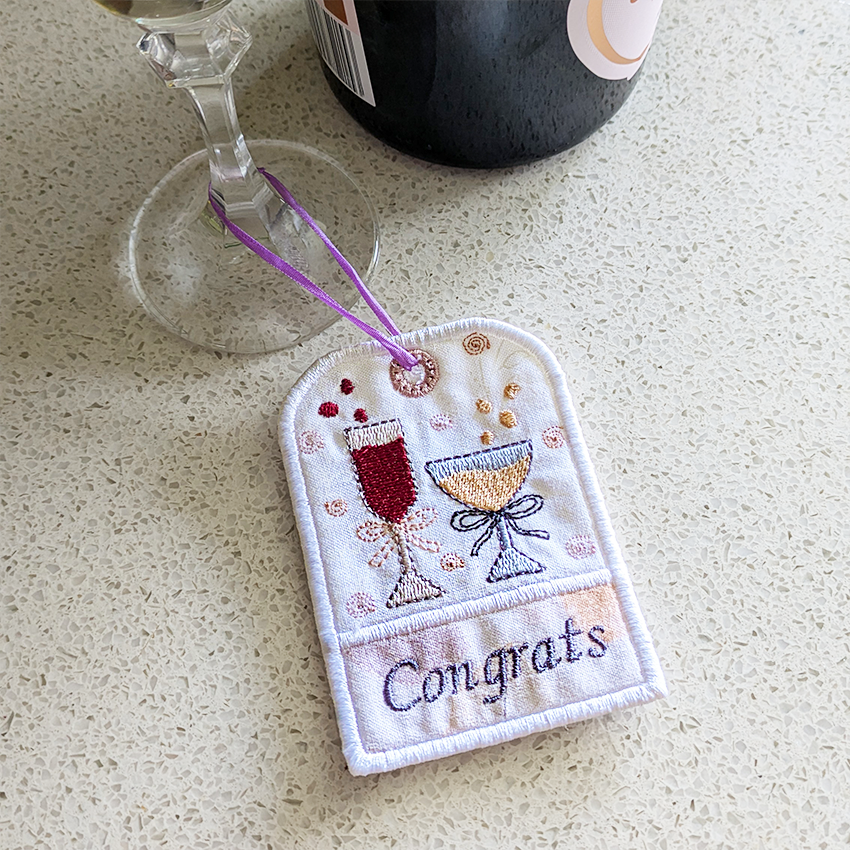 Embroidered 'Congrats' tag with wine glass and champagne flute design with a glass and bottle